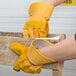 A person wearing yellow Cordova warehouse gloves with russet leather palms holding a wooden pallet.