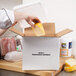 A hand placing a block of cheese in a white Nordic insulated shipping box.