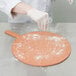 A person wearing white gloves using an American Metalcraft pizza peel to put flour on a pizza board.