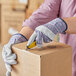 A person wearing Cordova striped canvas work gloves using a yellow and black box cutter to cut a box.