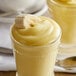A glass of yellow banana pudding with a white object on top and a banana on the side.