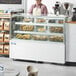 An Avantco bakery employee wearing an apron and standing behind a white dry bakery display case full of pastries.