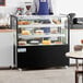 A person standing behind an Avantco refrigerated bakery display case with cakes on it.