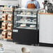 A man wearing an apron standing behind an Avantco refrigerated bakery display case with cakes in it.