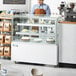 A man standing behind a white Avantco refrigerated bakery display case with cakes.