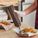 A brown 24 oz. wide mouth squeeze bottle being used to dispense sauce into a small container next to fried food in a basket.