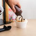 A hand holding a brown Choice wide mouth squeeze bottle pouring chocolate syrup into a cup of ice cream.