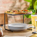 A black metal pizza stand holding a pizza on top with stacked white plates underneath.