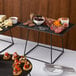 An Acopa black metal display stand holding a bowl of chocolate covered cherries on a table with nuts, fruits, and bread.