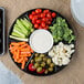 A Fineline black plastic 7-compartment tray with broccoli, baby carrots, tomatoes, and dip.