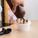 A hand holding a brown Choice wide mouth squeeze bottle pouring chocolate syrup into a cup of ice cream.