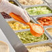 A hand holding a Carlisle orange measuring spoon over food.