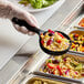 A person in gloves holding a Carlisle black acetal portion spoon over a bowl of food.