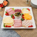 A white Fineline square plastic catering tray with meat, cheese, olives and tomatoes on a table.