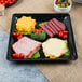 A Fineline black plastic square catering tray with cheese, meat, and vegetables on a table.