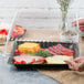 A hand holding a Fineline clear plastic container with a tray of food and a clear plastic lid.