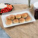 A Fineline white plastic rectangular cater tray filled with rolls next to a bowl of tomatoes.