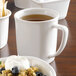 A white mug filled with brown liquid on a table with a bowl of cereal and blueberries.