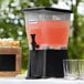 A black Choice 3 gallon beverage dispenser on a table with drinks and glasses.