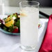 A Libbey clear frosted zombie glass filled with water and ice with a lemon slice on the rim on a table with a bowl of fruit and salad.