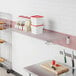 A Regency chrome wire wall mount shelf in a bakery display room with clear plastic containers with red lids on it.