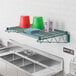 A Regency green epoxy wire wall mount shelf above a stainless steel sink with items on it.