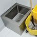 A stainless steel floor mounted mop sink with a mop and bucket with a handle.