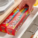 A hand using SC Johnson Saran Premium plastic wrap to cover a red box on a metal surface in a home kitchen.