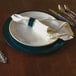 A blue Tabletop Classics by Walco plastic charger plate on a table with silverware and a napkin