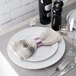 A purple Tabletop Classics napkin ring on a white plate with a knife and fork on a napkin on a table