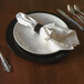 A Tabletop Classics black plastic charger plate with silverware and a white napkin on a table.