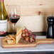 An American Metalcraft bamboo serving peel with a bottle and glass of wine next to a cutting board with cheese and grapes.