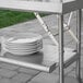 A Regency stainless steel folding work table with a stack of white plates on it.