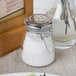 A Tablecraft glass salt shaker with a stainless steel clip-top lid on a table.