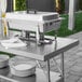 A Regency stainless steel folding work table with a chafing dish and stack of plates on it.