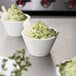 A white bowl of food with a sprig of thyme on top next to small white cups of green food.