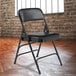 A National Public Seating black metal folding chair with a black vinyl padded seat in front of a brick wall.