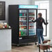 A woman standing in front of a black Avantco swing glass door refrigerator.