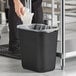 A person putting a plastic bag in a black Lavex rectangular trash can.