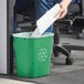 A person sitting at a desk puts paper in a Lavex green rectangular recycling wastebasket.