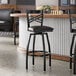 A black metal bar stool with a cross back design and a black vinyl padded swivel seat.