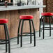A row of Lancaster Table & Seating black bar stools with red seats in front of a bar.