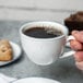 A person's hand holding a 10 Strawberry Street Arctic Blue porcelain cup of coffee with foam on a table.