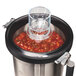 A Hamilton Beach stainless steel blender container on a table with a glass of tomato sauce inside.