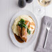 A Tuxton Charleston white china oval platter with mashed potatoes and sausages on it.