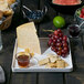 A white Arcoroc square porcelain plate with cheese, grapes, and fruit on a table.
