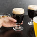 A hand holding a Libbey Banquet goblet filled with dark liquid on a table with another filled goblet and a bowl of nuts.