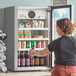 A woman opening the swing door of a Avantco countertop display refrigerator to take a beer.