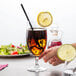 A hand holding a Chef & Sommelier iced tea glass of liquid with ice cubes and a lemon wedge.