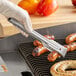 A person using Edlund heavy-duty gripper tongs to pick up sausages on a grill.